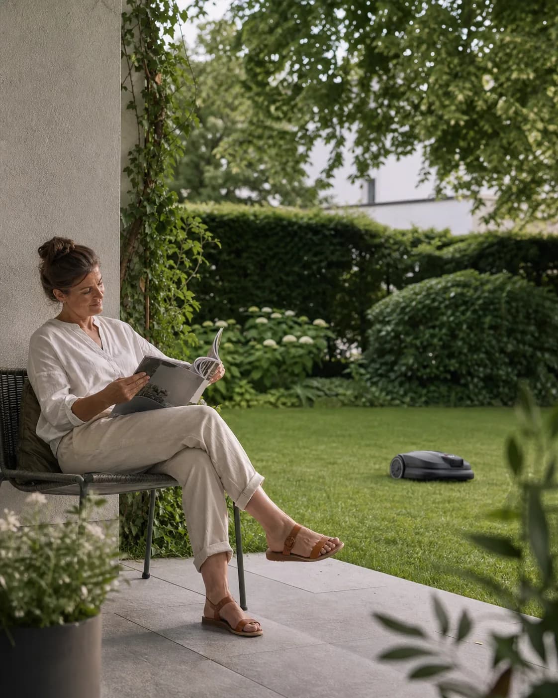 Frau liest auf der Terrasse während ein Mähroboter arbeitet